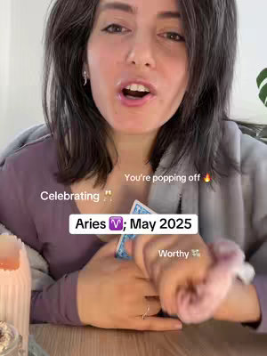 A woman with dark hair and fair skin is centered in the frame, looking directly at the camera with an open mouth, as if speaking. Text overlays: Aries V; May 2025, Celebrating and Worthy, arranged around her. Behind her, a soft grey-purple draped fabric is partially visible. She is holding a card, possibly a tarot card; the background has a soft, warm lighting.