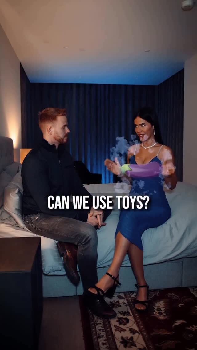 A man and woman are in a bedroom setting, seated on a bed. The woman is holding a toy shaped like an eggplant with vapor or steam emerging from the ends of the toy, as if in use. The text overlayed asks "Can we use toys?" The lighting suggests an intimate setting with a dark blue backdrop.