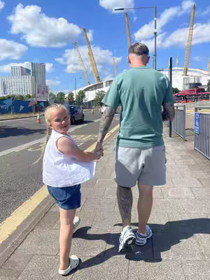 A sunny day scene captures a young girl and a man, possibly her father, holding hands as they walk on a sidewalk. The girl is wearing a white top and denim shorts, smiling at the camera. The man, seen from the back, has tattoos on his arm and is wearing a green t-shirt and gray shorts.
The background showcases a modern building and a large event structure under a blue sky with scattered clouds.