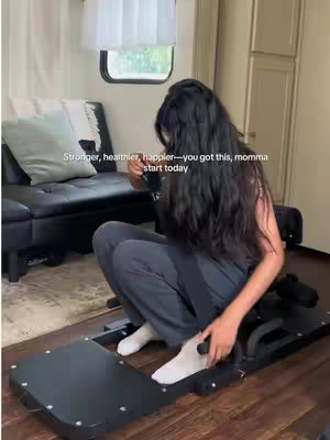 A woman with long, dark hair kneels on a black exercise machine in what appears to be the interior of a trailer or camper. She is dressed in dark grey athletic wear and white socks. Behind her is a black couch with a light blue pillow. Text overlay at the top says, "Stronger, healthier, happier — you got this, momma, start today."