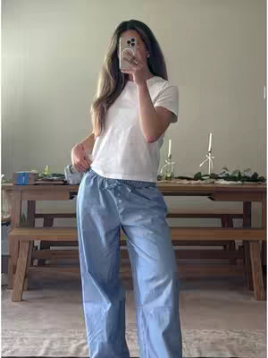 A woman stands, holding a phone in front of her face, obscuring it. She wears a white t-shirt and light blue pants. The background has a wooden table and a neutral-colored wall. Ambient lighting and basic decor suggest an interior setting.