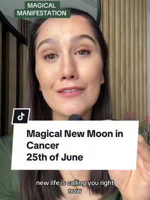 A woman with dark hair and a green shirt is centered in the frame and holding a white card. The card has the text "Magical New Moon in Cancer 25th of June" and beneath it reads "new life is calling you right now". Above her head in a green box reads "Magical Manifestation". The background is a light wooden slat wall. The image is well lit, likely a selfie shot.