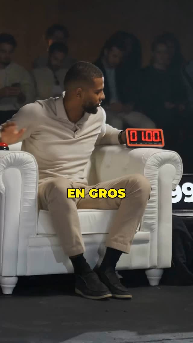 A man in a light-colored outfit sits in a white armchair in front of a dark background. A digital timer displaying 0:00 sits to the right of the man. The text overlay "EN GROS" in a yellow box appears below the man.