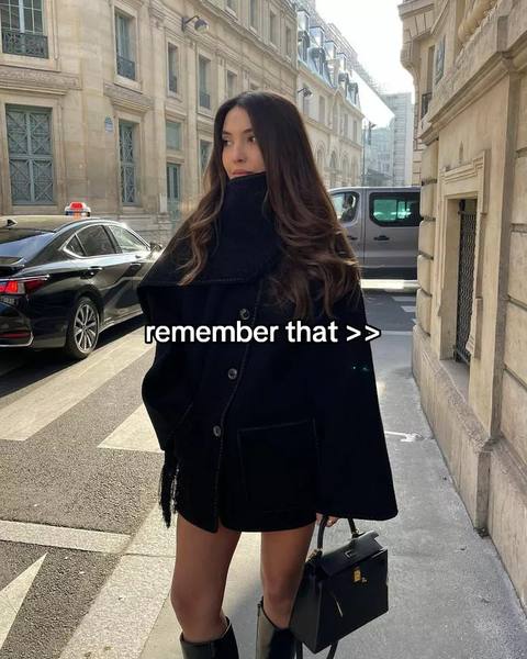 A woman in a black coat and boots stands on a city street in front of a blurred background with buildings and cars. Text overlayed on the image reads "remember that > >".