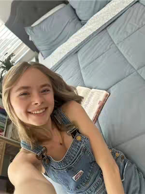 A young woman with long blonde hair is smiling while taking a selfie lying on a light blue comforter covered bed. She is wearing denim overall shorts and a necklace. An open book rests on her chest. The background features a grey headboard, pillows, and a cabinet.