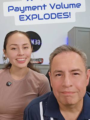 A selfie-style shot featuring a woman and a man smiling at the camera. Above them, a text overlay reads, "Payment Volume EXPLODES!" in blue and white. The woman is on the left, and wears a pink shirt next to the man wearing a dark blue top. They are positioned in front of a light-colored wall with a blurred logo.