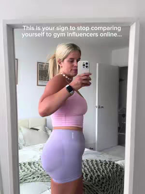A woman takes a selfie in a mirror, showcasing her body in workout clothes. She wears a pink sports bra and light purple shorts, with her hair in a ponytail. Text overlays read, 'This is your sign to stop comparing yourself to gym influencers online...' Framing includes a mirror, a wall, and a glimpse of a bed.