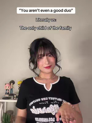 A woman with dark hair and bangs stands in front of a neutral-colored wall. She wears a black t-shirt with a white collar and a graphic design. The text overlay at the top says: 'You aren't even a good duo'. Text below reads: 'Literally us: The only child of the family'. Miniature figures are arranged on a shelf behind her.
