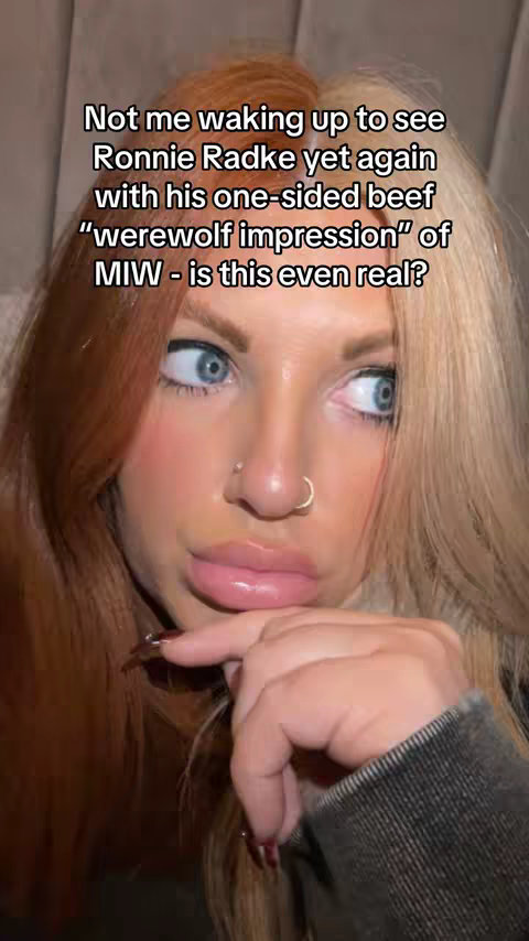 A close-up selfie of a person with striking features and a two-toned hair style. The individual has fair skin, full, glossy lips, and blue eyes. The hair is parted to the side, with one side a bright auburn and the other a light blonde. The person is wearing a dark gray or black denim jacket, and a small nose ring is visible. The background is blurred, suggesting a soft focus, and the overall composition is close and personal.