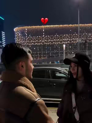 In an outdoor setting at night, a man and a woman face each other, with a red, broken heart graphic overlayed above them. The man is to the left, wearing a coat with a fur collar, while the woman is on the right, wearing a cap. In the background, a modern building with illuminated details is visible, along with the blurred lights of the city and a car.