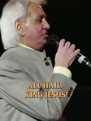 A medium shot of a man speaking into a microphone. The man, likely in his 60s or 70s, has short, slicked-back white hair and wears a light gray suit with a yellow collared shirt. The microphone is black and silver. Overlayed on the image is text.