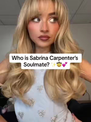A selfie-style image featuring a woman with long blonde hair, bangs, and a light blue or silver embellished top. She gazes to the side with a thoughtful expression, and a text overlay reads, 'Who is Sabrina Carpenter's Soulmate?' with a cowboy and heart emojis.