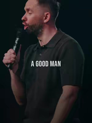 A man with short dark hair and a trimmed beard is holding a microphone and talking. A text overlay in white letters reads 'A GOOD MAN'. The background is dark and blurry.