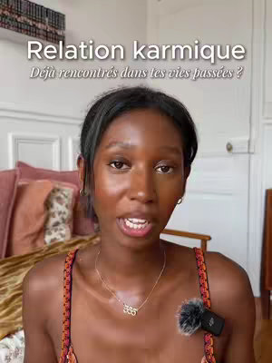 A woman with dark hair and skin is centered in the frame. She is wearing an embellished top and a gold necklace with the number 333 on it. A microphone is attached to her shirt. In the background, there are pillows and a bed. Text overlayed on the top says "Relation karmique" and "Déjà rencontrés dans tes vies passées ?".