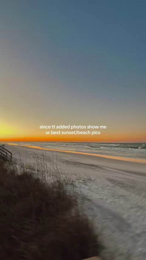 The image captures a serene beach scene at sunset. The sky is a gradient of colors, transitioning from a deep blue at the top to vibrant oranges and yellows near the horizon, where the sun is setting. Below the sky, the ocean meets a sandy beach, and the foreground is composed of grassy dunes and a wooden walkway leading to the beach. The composition uses natural elements with a warm color palette.