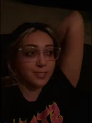 The image shows a woman with blonde-streaked hair, wearing glasses and a black t-shirt with a graphic, likely posing, and resting her arm above her head. The background is dark, creating a silhouette effect. The lighting is soft and warm, focused on the woman's face.