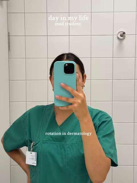 A selfie taken in a white-tiled restroom. The person, whose face is mostly covered by a turquoise phone, wears a green medical scrub. Text overlays are visible. The overall composition is simple, with a focus on the mirror reflection.