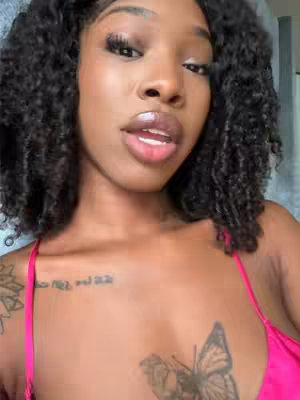 A close-up shot of a woman with curly hair and a pink top. She has tattoos on her chest and arm. The background is blurred, but appears to be an indoor setting.