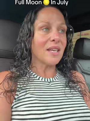 A woman with curly dark hair is seated in a car, looking slightly to her right and speaking. She has light skin and is wearing a black and white striped tank top. Visible is a car interior with a black leather seat and the window.