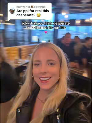 A smiling young woman with long blonde hair is positioned in the foreground, set against a blurred background of a bar or restaurant. She wears a black leather jacket and small hoop earrings, and her expression is warm and engaging, with a slight smile. The background features indistinct figures and warm lighting, suggesting a social setting.