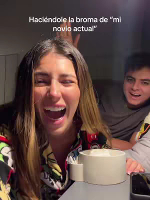 A woman is laughing with her mouth wide open, and a man is smiling behind her. Above them, text reads 'Haciéndole la broma de "mi novio actual".' To the right of the woman, a small white container with a handle sits on a surface.