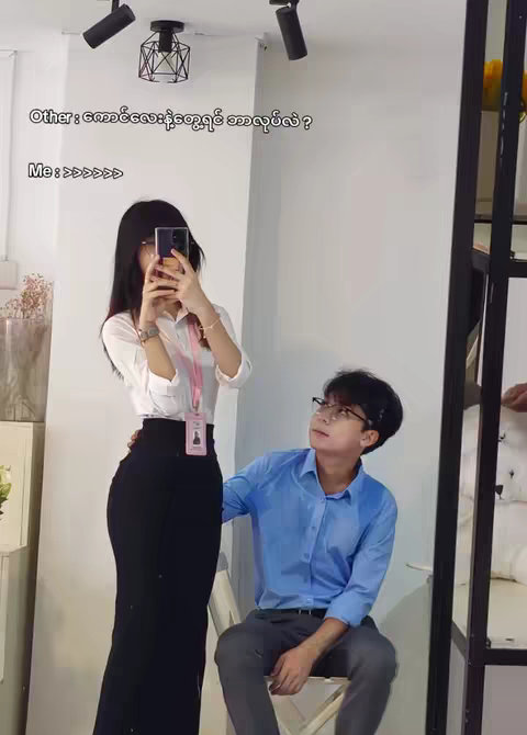 A photo taken indoors shows a woman in a white shirt and black skirt, holding a phone up to take a selfie, facing a man who is looking up at her. The woman has her long black hair parted in the middle and her face partially obscured by the phone. The man is wearing a blue button-down shirt, glasses, and is seated on a chair. The background includes a white wall, a mirror, and some lighting fixtures.