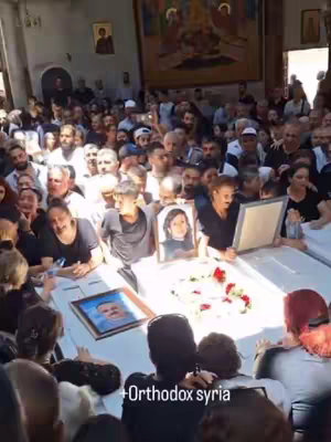 The image shows a large crowd of people gathered inside what appears to be a church or a similar religious building. The focus is on a central area where several individuals are holding portraits of deceased people, likely in memory of those who have passed away. In the background, there's a large painting depicting religious figures and themes. The overall atmosphere seems somber and mournful, reflecting a time of bereavement. The color palette is natural, dominated by the clothing. The lighting is mixed, using both natural and artificial sources.