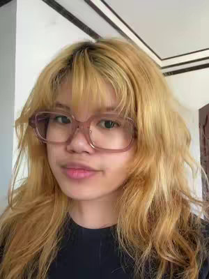 A selfie-style shot of a person with fair skin, wearing large, light-brown framed glasses. They have blond hair with bangs and are wearing a dark-colored top. The background is a plain wall and ceiling.