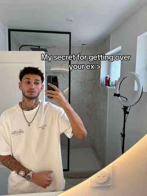 A man takes a selfie in a bathroom with a large illuminated ring light. The man is wearing a white t-shirt and has multiple tattoos. Text overlay reading, “My secret for getting over your ex >” is on the mirror.
