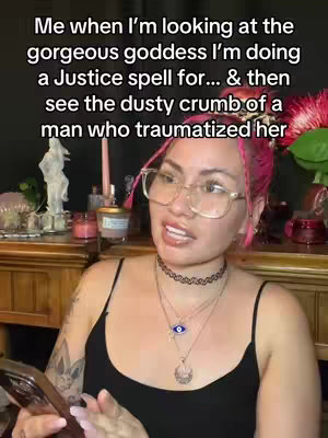 A woman with pink-streaked hair, wearing glasses and multiple necklaces, is positioned in front of a dimly lit scene. Text overlays the image, and she appears to be talking, with a phone in her hand.