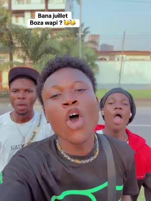 Three young men are captured in a selfie-style shot. The central figure is close to the camera, with an open mouth suggesting they are speaking or singing. The individuals stand in front of outdoor features, some trees and buildings are also visible in the background. The overall image conveys a casual and energetic vibe, likely of a social media post.