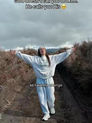 A woman stands outdoors with arms outstretched, framed against a cloudy sky and brown background. She wears a white sweatshirt and light-colored pants. The image suggests a feeling of freedom and spiritual connection.