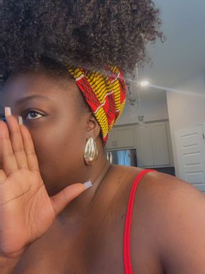 A close-up of a Black woman covering her mouth with her right hand. She has curly hair pulled back with a patterned headscarf with red, yellow, and black elements. She wears large gold hoop earrings and a red top, with her left eye visible. The background has light-colored cabinets.