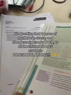 The image shows a close-up shot of a desk covered in papers and documents, with a handwritten text overlay. The papers are predominantly white with some colored markings and annotations. A pen rests on top of the papers. The background appears blurry, suggesting an indoor setting. The text overlay is prominent, featuring large, bold text against the cluttered background.