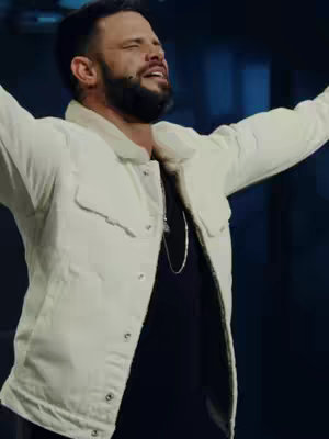 A medium shot depicts a man with short dark hair and a well-groomed beard, gazing upwards with his arms outstretched in a gesture of worship and reverence. He wears a white jean jacket with a shearling collar over a black top. The background is blurred, with a blue hue and suggests a stage setting.