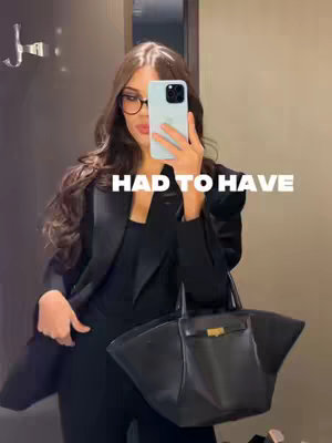 A mirror selfie featuring a woman in a dark suit, holding a light blue phone in front of her face, obscuring the face. She wears glasses and has long, wavy, dark hair. A black bag is held in front of her, with a light reflection from the mirror. The background is a nondescript space, likely a hotel room or similar setting.