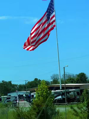 A photograph of an American flag waving in the wind. The flag is in the distance, attached to a tall pole. The sky is clear blue. In the foreground are green trees and bushes and some buildings, RVs and parking spaces are visible in the background. The image is well-lit, with bright light.