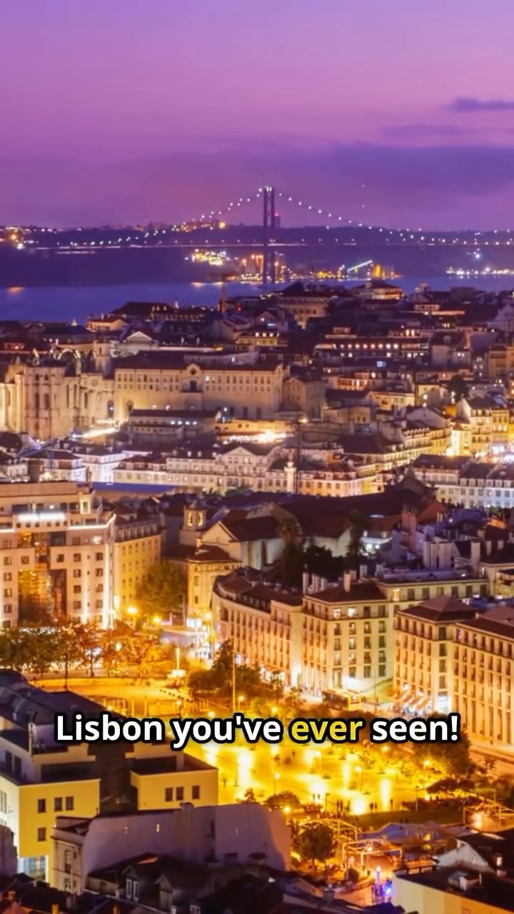 An aerial view of Lisbon, Portugal at dusk or dawn. The city is densely packed with buildings, and a bridge is visible in the distance.