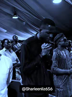 A low-angle shot inside a well-lit space, likely a church or auditorium. A crowd of people are present, dressed in semi-formal attire. The focus is on a man in the foreground; he is dressed in a black shirt, his hands clasped in prayer, with eyes closed. The overall tone is reverent, and the lighting gives the scene a slightly cool color cast.