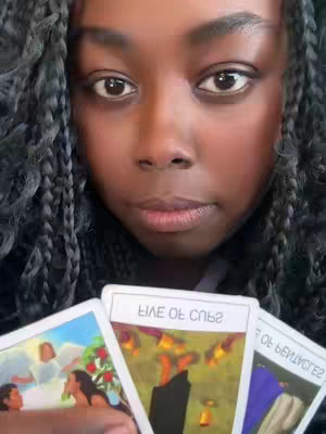 A close-up shot featuring a woman with braided hair, looking directly at the camera with a neutral expression. She holds three tarot cards, partially obscuring her lower face. The cards are oriented toward the viewer and display various illustrations and text, suggesting a reading or divination theme. The background is simple, focusing attention on the woman and the tarot cards.