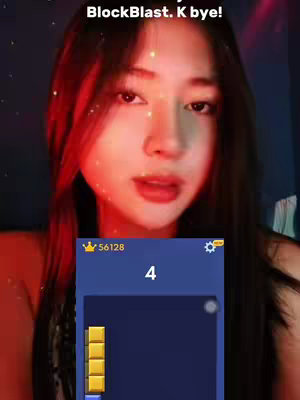 A young woman with long black hair is shown in a close-up shot. She's looking slightly towards the camera, with a focused expression on her face. A mobile game interface is superimposed near her chin, with the number '4' prominently displayed, and a game piece block below, creating a game context. Above her head is text and some glittery effects.