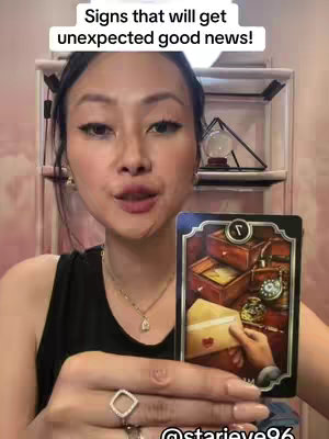 A woman with long dark hair is centered holding a tarot card featuring an image of a hand opening a letter from a box, surrounded by other boxes, with a decorative frame. Text at the top reads 'Signs that will get unexpected good news!'. The woman looks directly at the camera with a slight smile, her hands subtly positioned. The background is a light pink wall with decorative elements.
