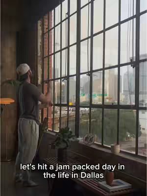 A man stands near a large window with numerous divided panes, looking out at a city skyline. The window frame is dark and contrasts with the brightness of the view. The man is wearing casual attire and a cap. Text, in a white font, appears at the bottom of the frame.