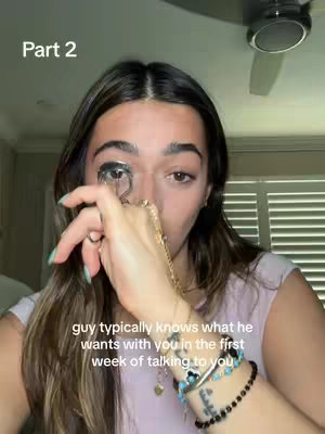 A selfie-style video showing a woman with dark hair holding an eyelash curler. The background is slightly blurred, suggesting an indoor setting with white blinds and a ceiling fan.