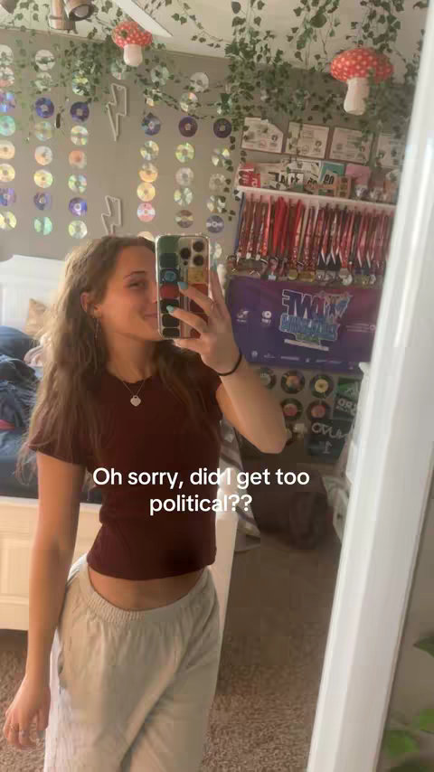 A selfie taken in a bedroom with a mirror capturing the reflection. The creator, a young woman, holds the phone up, partially obscuring her face. The background includes a bed, decorations, and a collection of medals.
