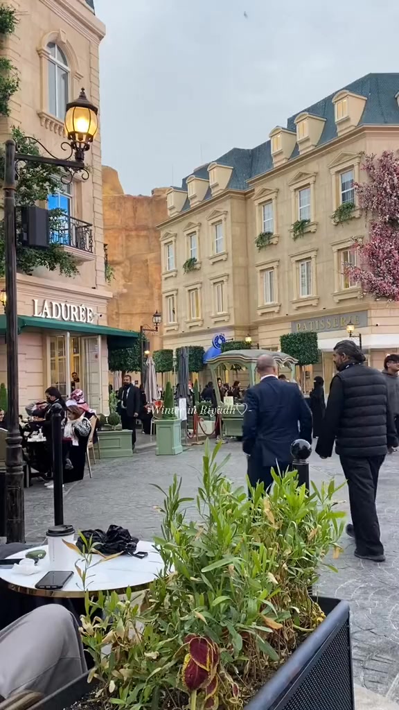 An exterior shot of a city street resembling a European town square. Buildings with light-colored walls, arched windows, and decorative details line the sides. Restaurants and shops with awnings offering outdoor dining. There are people walking around the square.