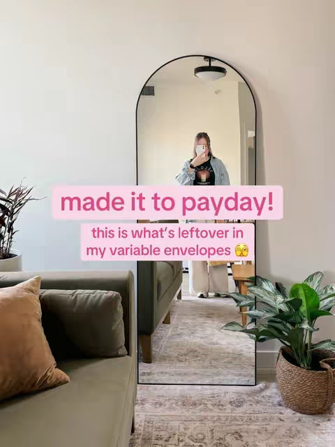 A person in a blue jean jacket and a black t-shirt holding a phone up in front of their face in a full-length arched mirror. The mirror reflects the persons surroundings, showing part of a green sofa, a rug, and a plant. White text inside a pink box overlaid on the image reads "made it to payday!" and "this is what's leftover in my variable envelopes 😳 ".