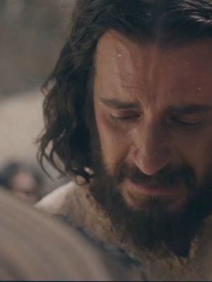 Close-up shot of a man with a beard and long dark hair, face tilted down, eyes closed as if in prayer or sorrow. The lighting emphasizes the man's features, with a focus on his expression.