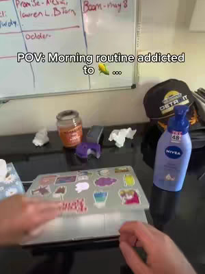 The image shows a desk cluttered with items including a laptop covered in stickers, skincare products, and various other items. The text overlay reads 'POV: Morning routine addicted to ...' .