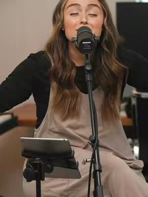 A woman with long brown hair is singing into a microphone. She is wearing a black long-sleeved shirt under a beige overall. The background is plain.